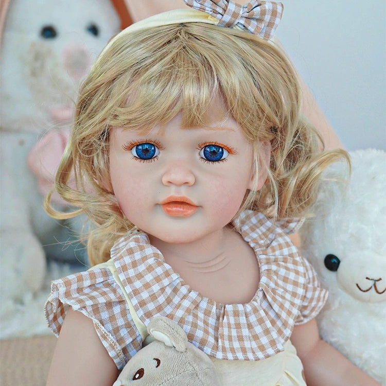 Reborn toddler doll sitting in a soft chair, dressed in white bunny-themed overalls and a pink top, holding a small plush toy.