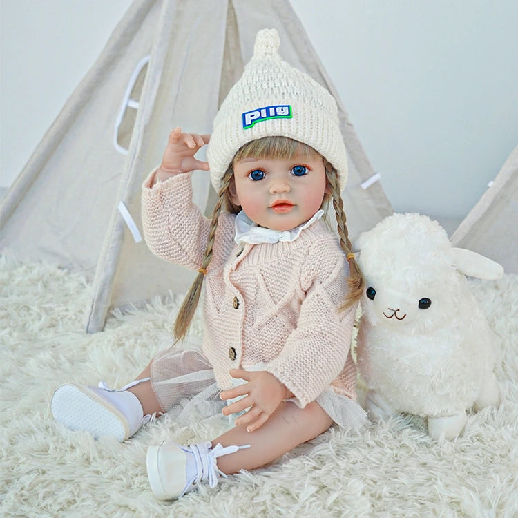 Reborn toddler doll sitting next to a stuffed animal, wearing a pink outfit and knit hat.