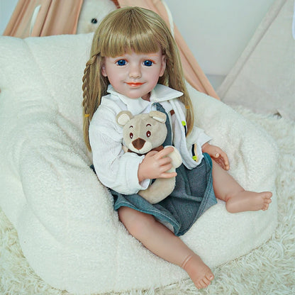 Reborn toddler doll sitting in a plush chair, dressed in a school-themed outfit with a teddy bear on her lap.