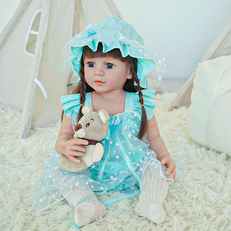 Reborn toddler doll sitting on a soft surface, dressed in a light blue polka dot dress, holding a small plush bear.