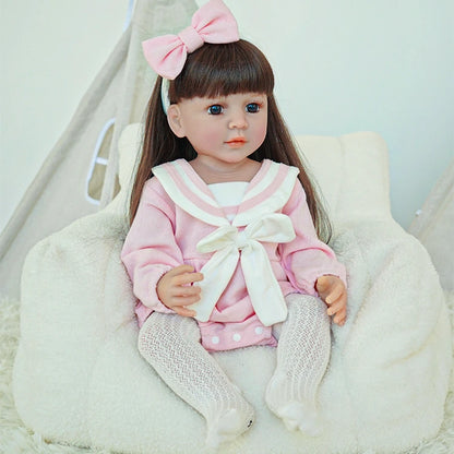 Reborn toddler doll sitting on a soft surface, dressed in pink with a white ribbon, wearing gray tights, and looking straight ahead.