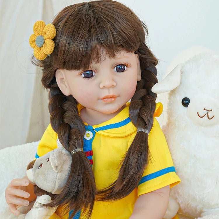 Reborn toddler doll sitting on a rug, dressed in a bright yellow and blue dress with white socks, holding a small teddy bear.