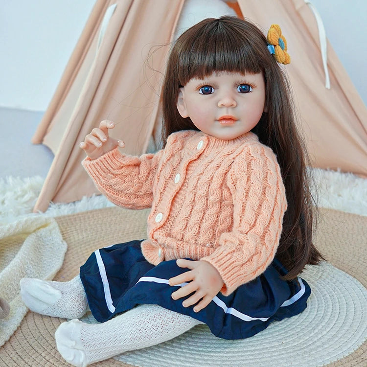 Reborn toddler doll sitting on a soft rug, wearing a peach knit sweater and navy skirt, looking forward.