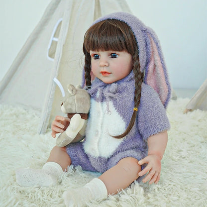 Reborn toddler doll in a cozy purple bunny outfit, sitting on a fluffy rug with her hands resting in her lap.