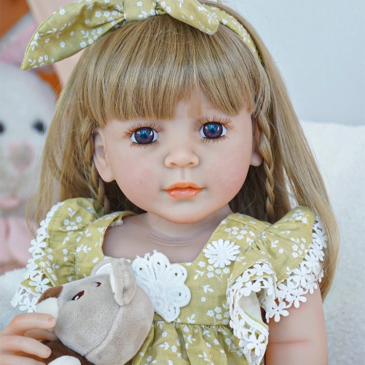 Reborn toddler doll sitting on a soft rug, wearing a green floral dress with white socks, holding a teddy bear, looking to the side.