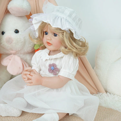 Reborn toddler doll sitting with her hands together, wearing a white dress with a bonnet, looking gently to the side.