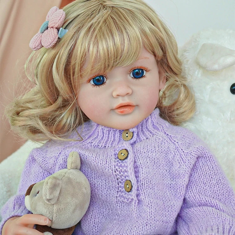 Close-up of reborn toddler doll wearing a purple sweater and plaid pants, holding a plush bear, with soft curls and a pink hair clip.