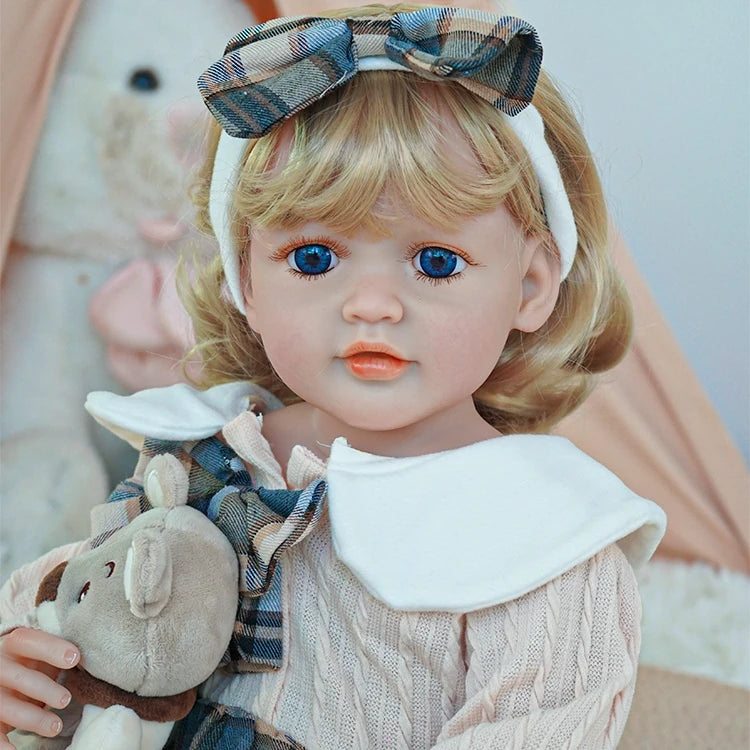 Close-up of reborn toddler doll with soft blonde curls and a plaid bow headband, dressed in a plaid dress, holding a teddy bear.