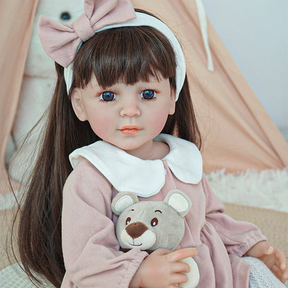 Reborn toddler doll with long straight brown hair and a pink bow, sitting on a rug with her plush toy.