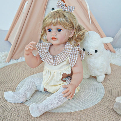  Reborn toddler doll sitting in a plush chair, wearing white overalls with a bunny design, holding a teddy bear.