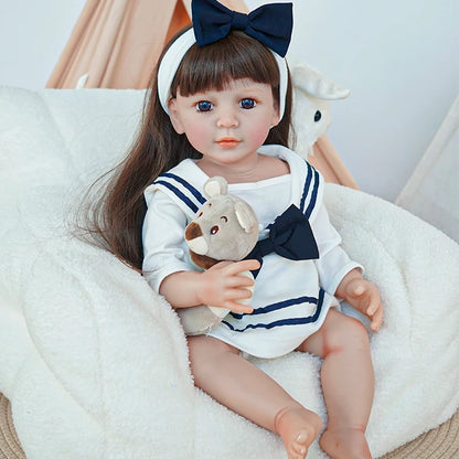 Reborn toddler doll sitting on a chair, wearing a navy and white sailor outfit, holding a teddy bear, with a navy blue bow on her head.