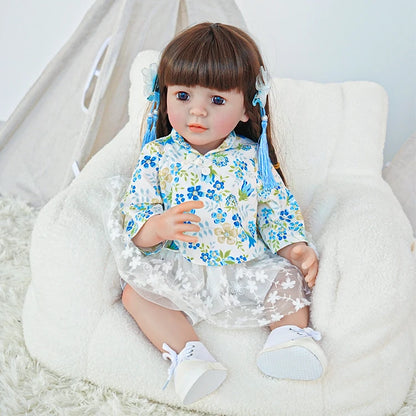 Reborn toddler doll sitting in a plush chair, dressed in a blue floral top and white skirt, looking forward.