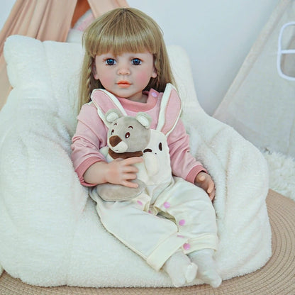 Reborn toddler doll sitting in a plush chair, wearing white overalls with a bunny design, holding a teddy bear.