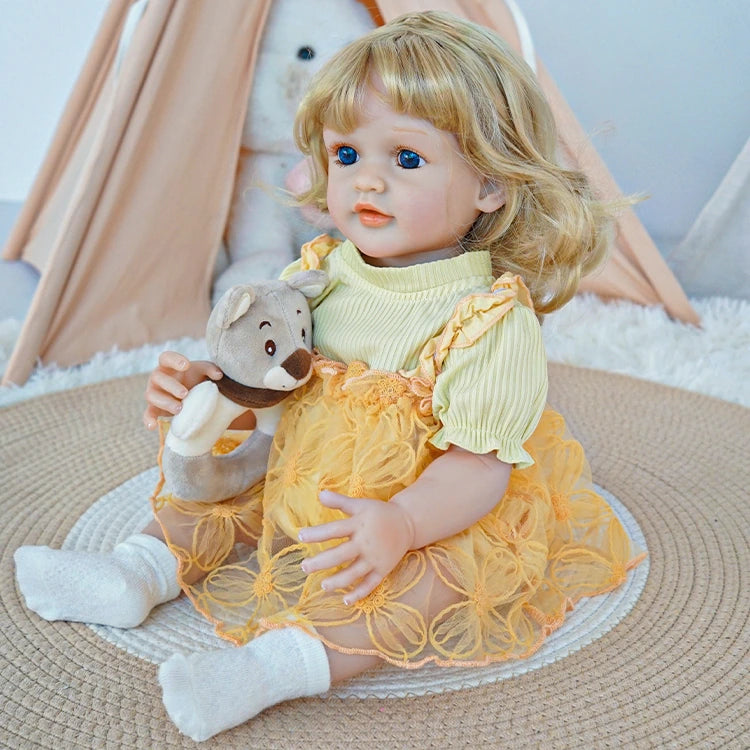 Reborn toddler doll sitting on a rug, dressed in a yellow floral dress, holding a small teddy bear, looking to the side.