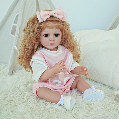 Reborn toddler doll holding a teddy bear, dressed in a pink dress with a bow, sitting on a soft blanket.