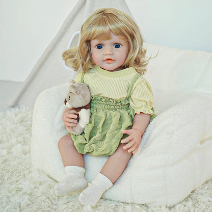 Reborn toddler doll sitting on a soft rug, dressed in a green dress and white socks, looking forward with a gentle expression.