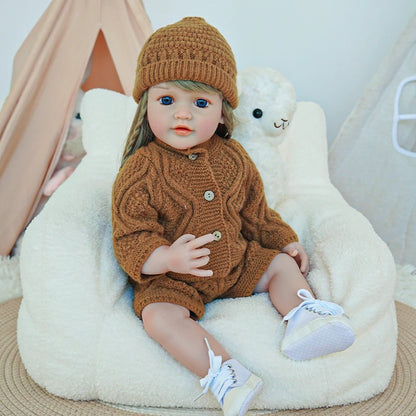 Reborn toddler doll sitting on a soft chair, dressed in a brown knit sweater and hat, holding a plush toy.