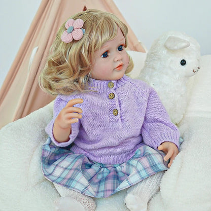 Reborn toddler doll sitting in a plush chair, dressed in a purple sweater and plaid pants, looking to the side.