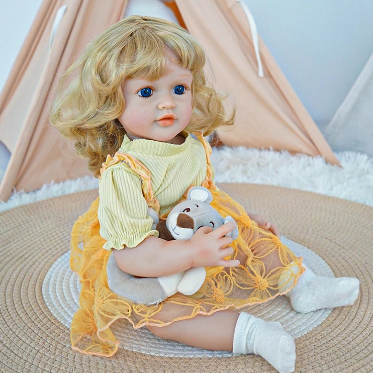 Reborn toddler doll sitting on a soft rug, dressed in a yellow dress with floral details and white socks, holding a teddy bear.