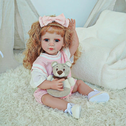 Reborn toddler doll sitting on a plush surface with her teddy bear, styled with a pink dress and matching hair bow.