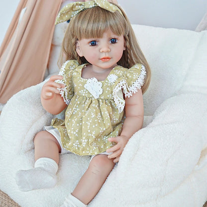 Reborn toddler doll sitting on a plush surface, dressed in a green dress with lace accents, holding a small teddy bear, looking gently forward.