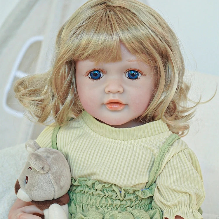 Reborn toddler doll sitting in a plush chair, wearing a green dress and holding a small teddy bear.