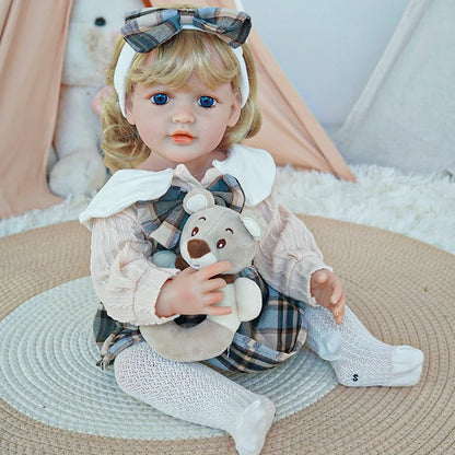 Reborn toddler doll sitting on a soft rug, dressed in a plaid dress with a white collar and white tights, holding a small teddy bear.