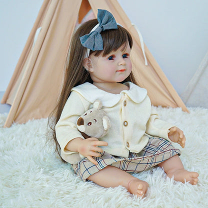 Reborn toddler doll sitting on a rug, wearing a plaid skirt and a cream top with a blue bow in her hair, holding a plush bear.