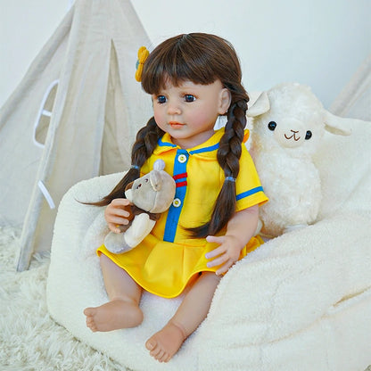  Reborn toddler doll sitting on a soft chair, wearing a yellow dress with blue accents and a flower in her hair, holding a plush toy.