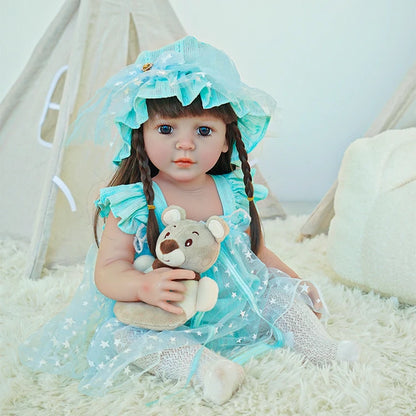 Reborn toddler doll sitting on a rug, dressed in a blue dress with a matching bonnet, holding a teddy bear.