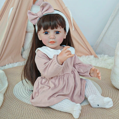 Reborn toddler doll sitting on a soft surface, dressed in a pink dress with a white collar and holding a small teddy bear.