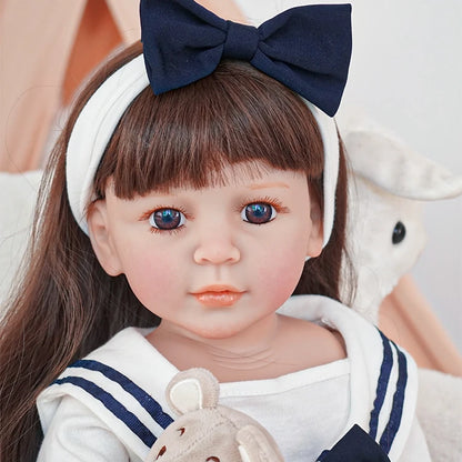 Reborn toddler doll wearing a sailor-style dress, looking gently forward while holding a teddy bear, with a large navy bow in her hair.