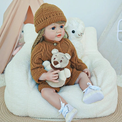 Reborn toddler doll wearing a cozy brown outfit and knit hat, sitting on a plush surface with a stuffed bear.