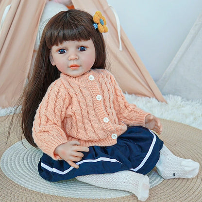 Reborn toddler doll sitting on a soft surface, dressed in a peach knit sweater and navy blue skirt, with white socks.