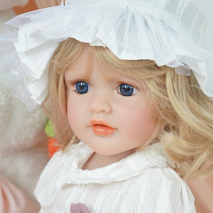 Close-up of reborn toddler doll wearing a white bonnet and dress, with soft curls framing her face, looking forward.