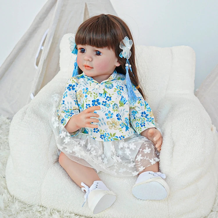 Reborn toddler doll sitting in a chair, wearing a floral blue top and skirt with a flower hair clip.