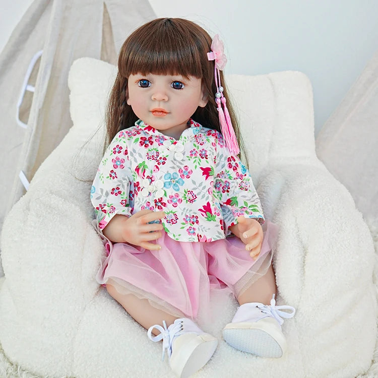 Reborn toddler doll sitting on a soft chair, wearing a floral pink top and pink skirt, holding a small teddy bear.