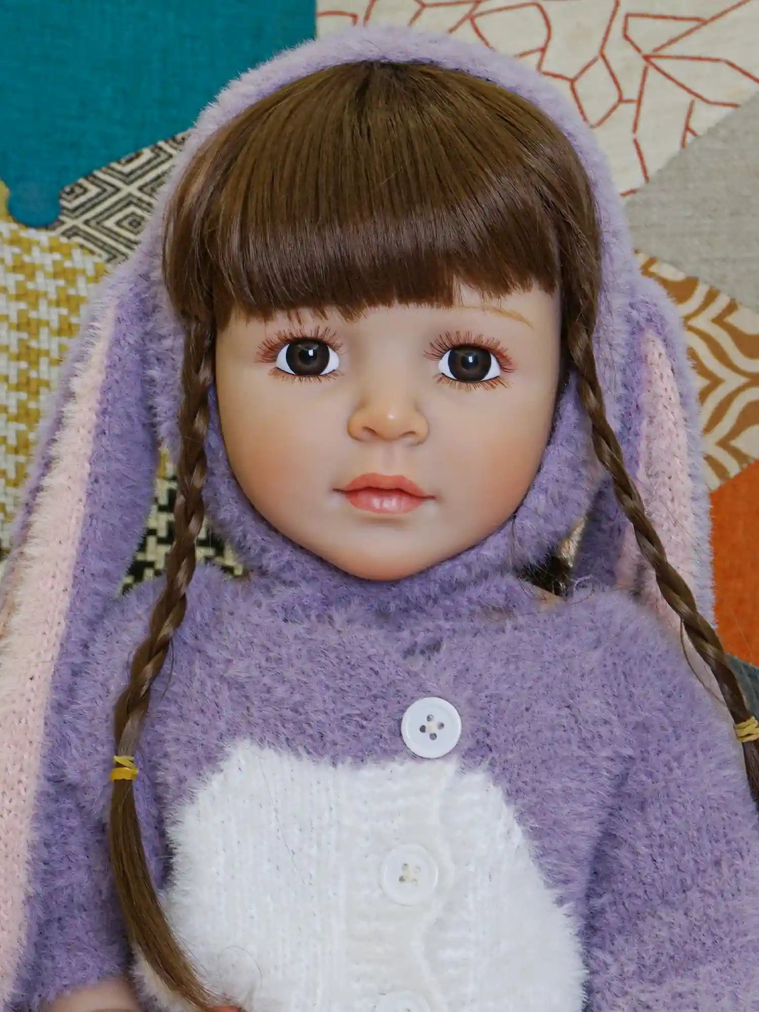 A sitting doll with a realistic face, brown hair in braids, and dressed in a textured purple bunny suit, surrounded by a learning clock and white flowers.