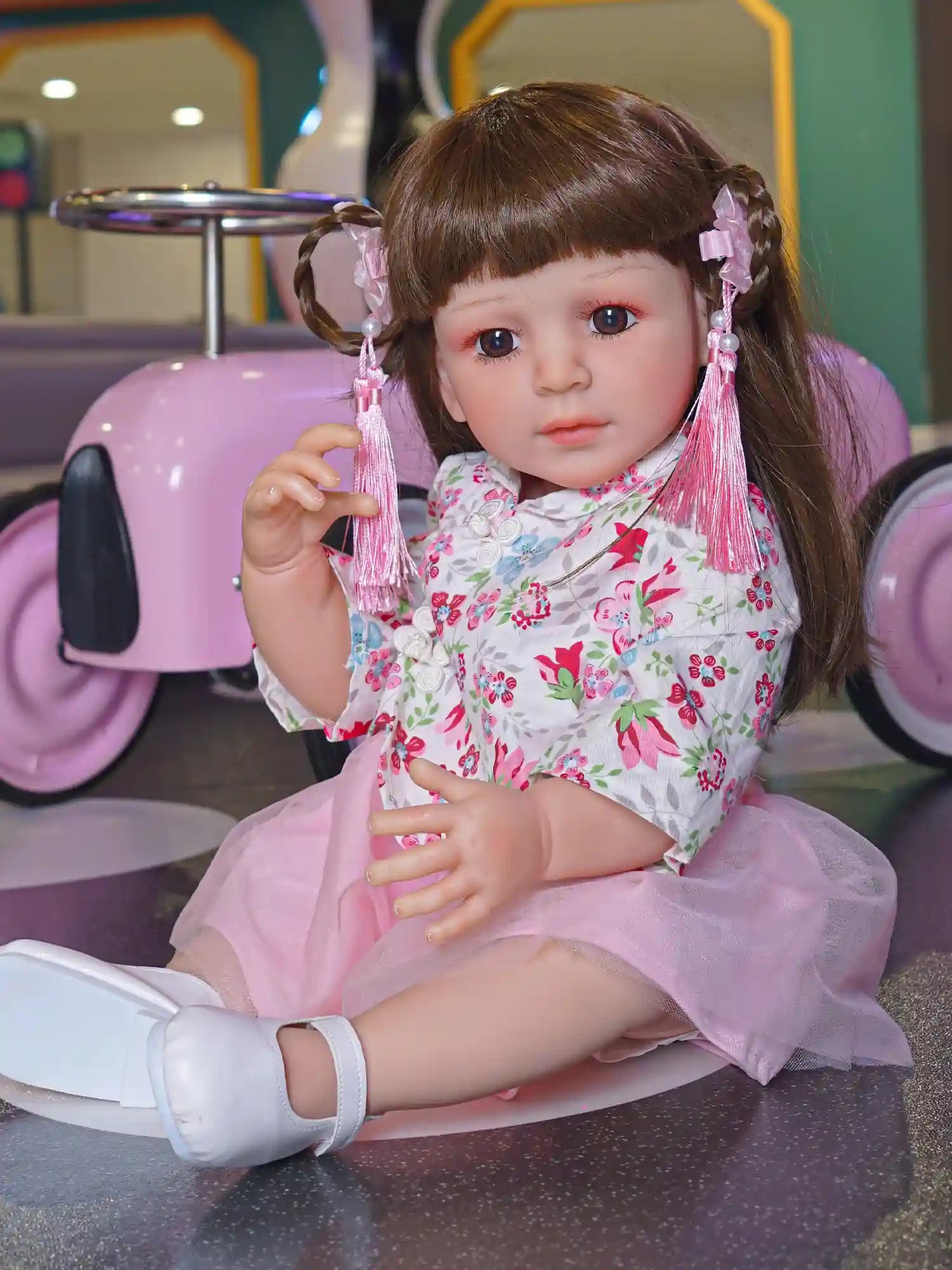 Close-up of a toddler doll in a pink and white floral kimono dress with pink ribbon hair accessories, posed playfully.