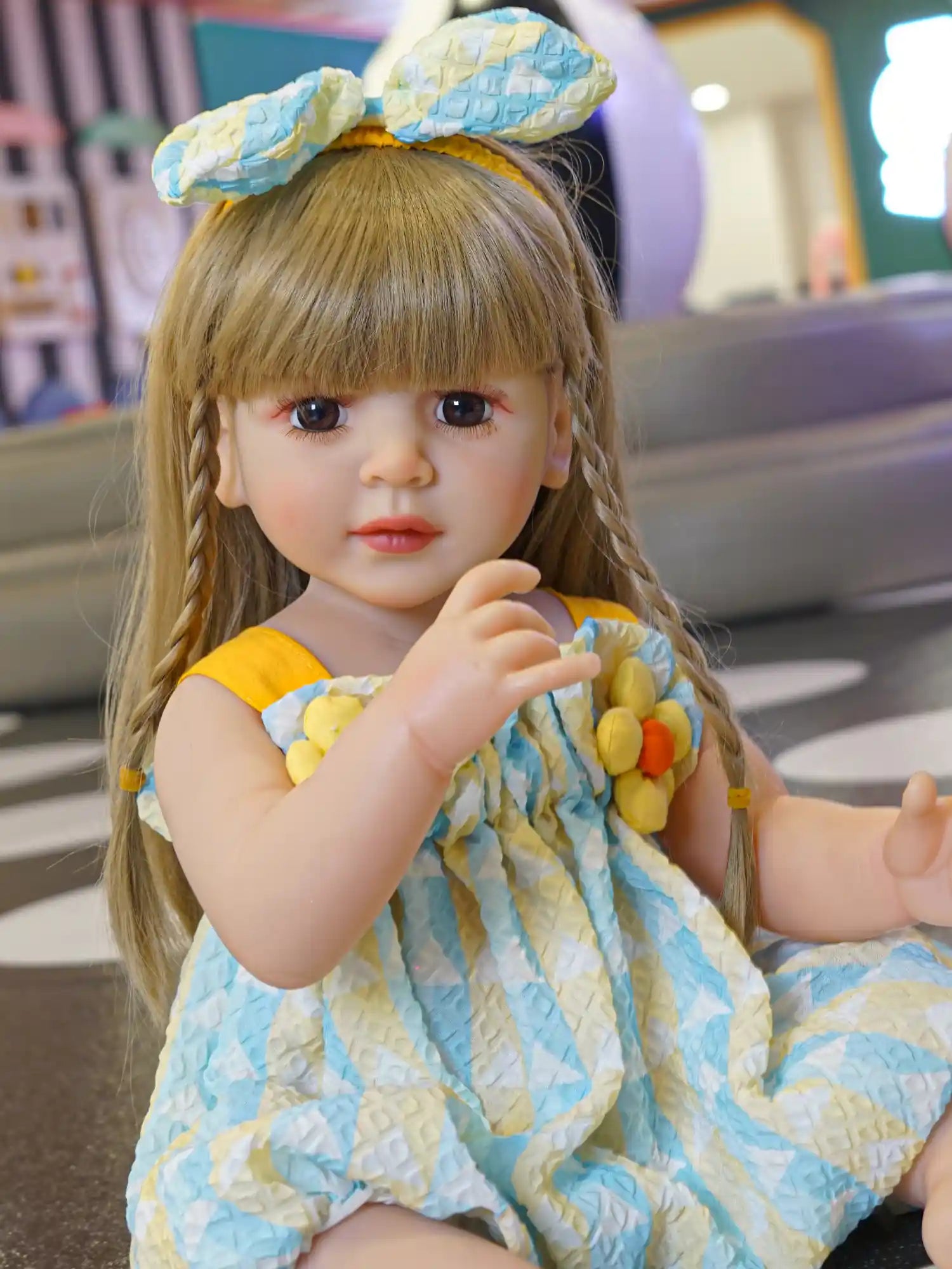 Lifelike blonde doll sitting in a blue and yellow dress with floral accents.