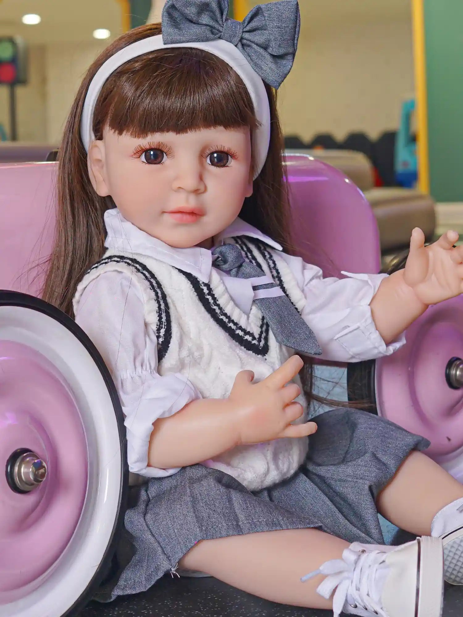 A doll with the likeness of a child at school, complete with brown hair and pensive brown eyes, dressed in a grey and white outfit and sitting by a pink toy car.