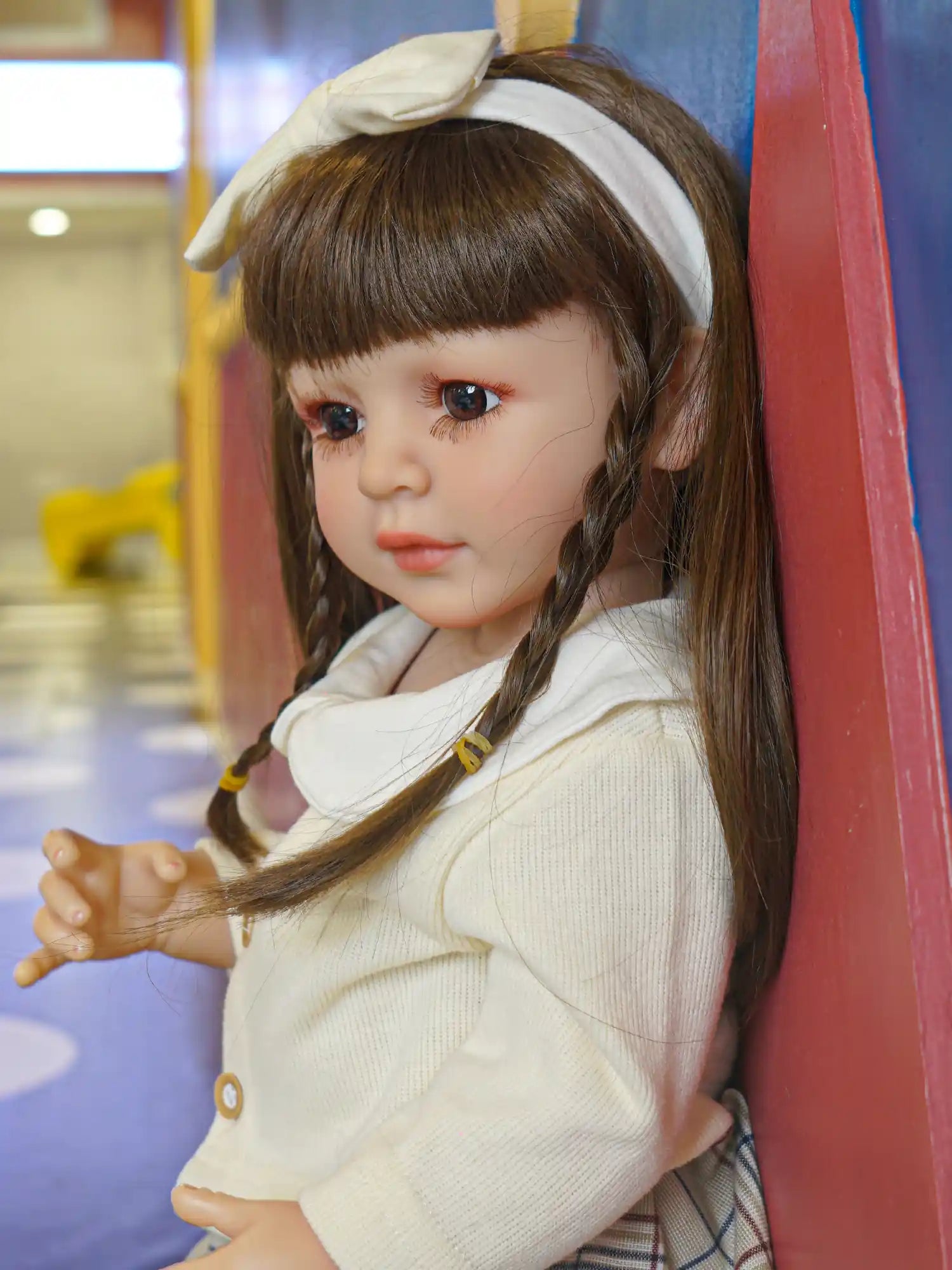 Doll with delicate facial features, wearing a white knitted sweater and a tartan skirt, seated on a shiny floor.
