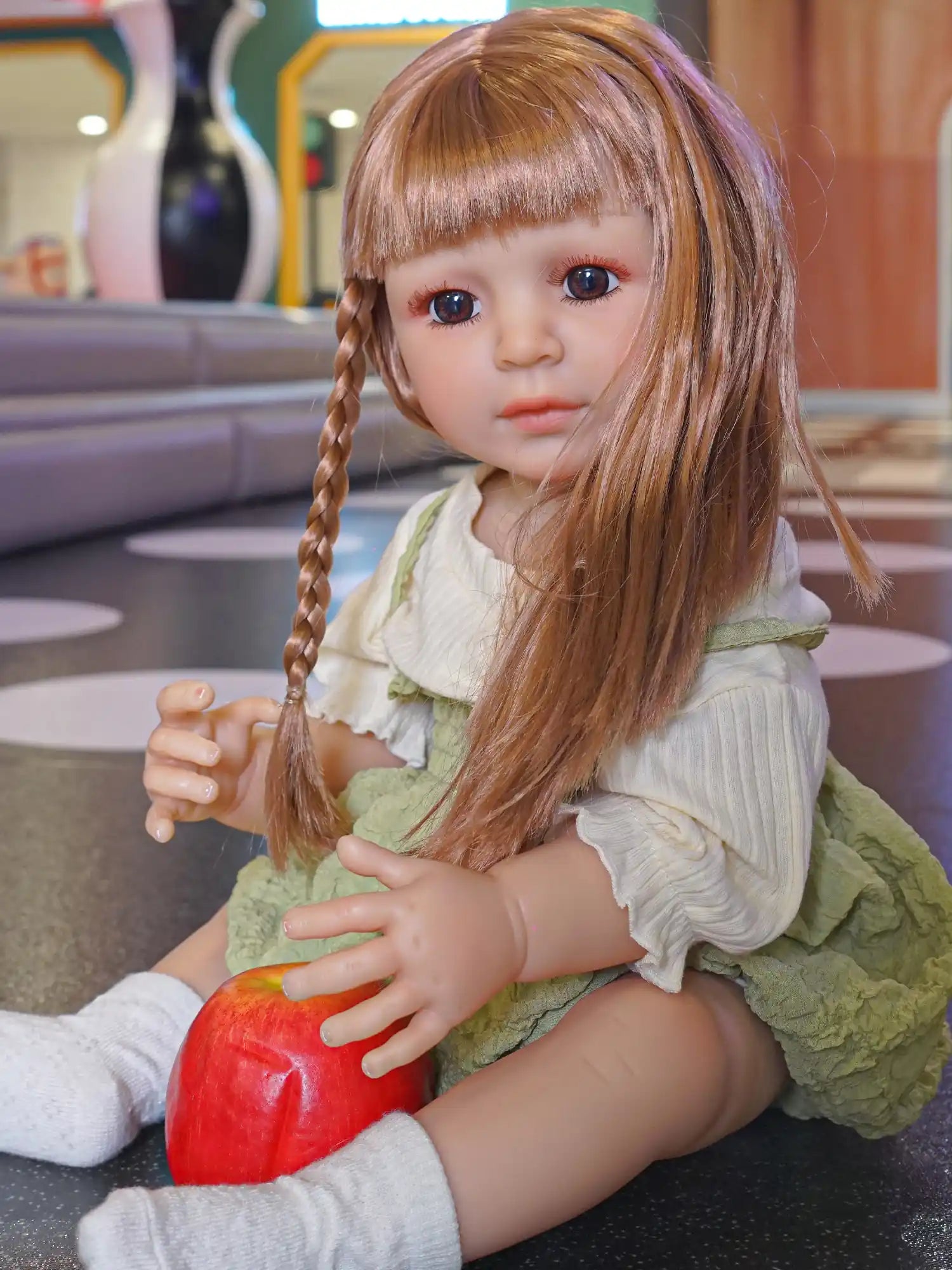 Doll with braids and green dress holding an apple, sitting on the floor.