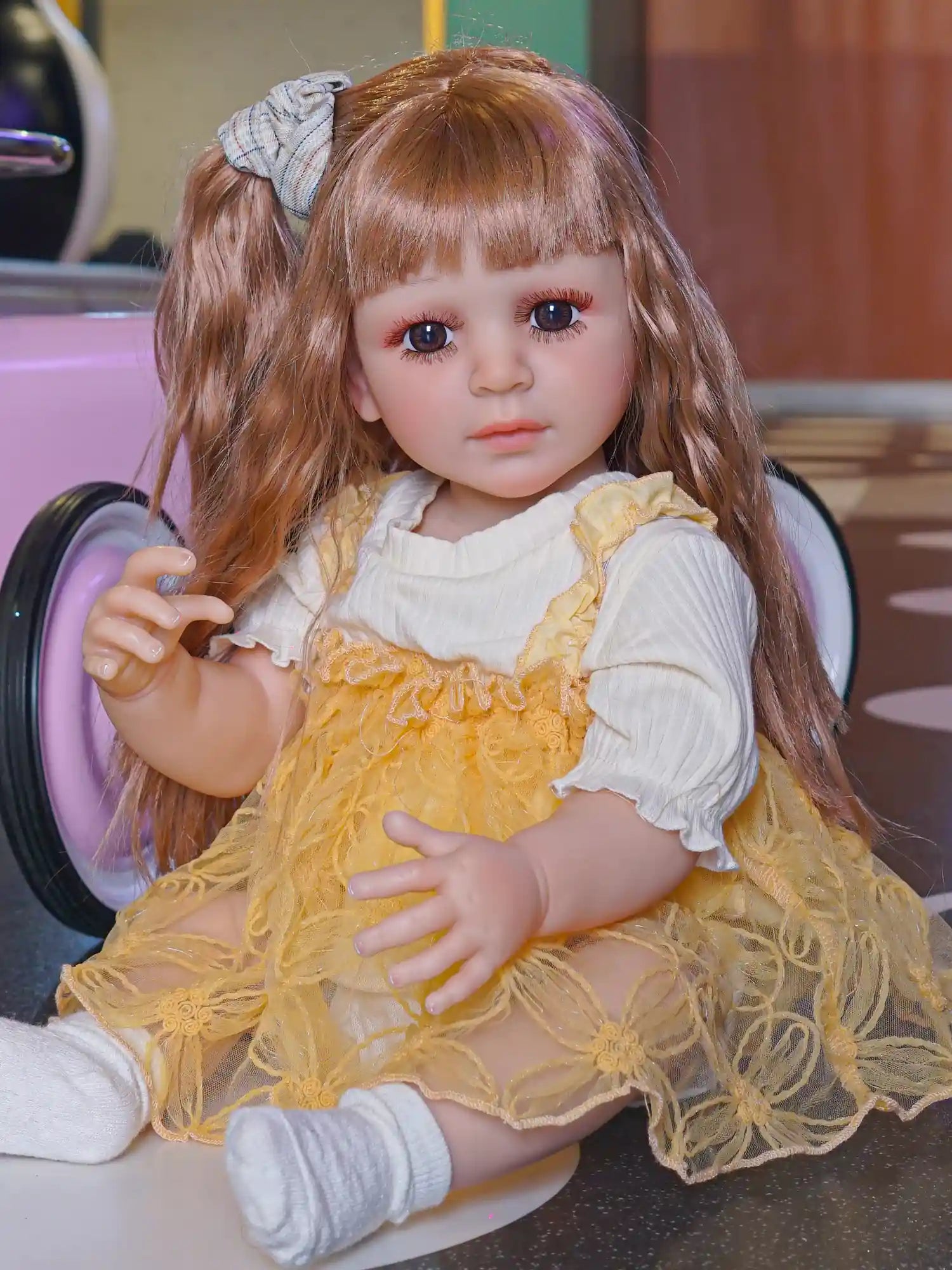 Toddler doll with wavy auburn hair and a plaid bow, wearing a yellow lace dress over a white blouse, seated with a toy car in the background.