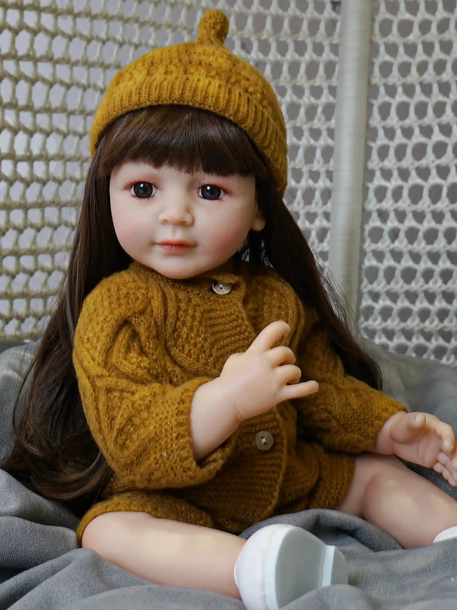 A doll with a realistic childlike demeanor, donning a mustard knit hat and sweater, capturing a relaxed pose on a comfortable gray textile.