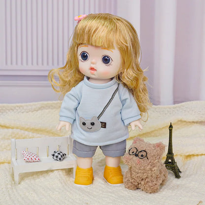 Doll with blonde curly hair and big blue eyes, dressed in a blue sweatshirt and grey shorts, accompanied by a plush toy.