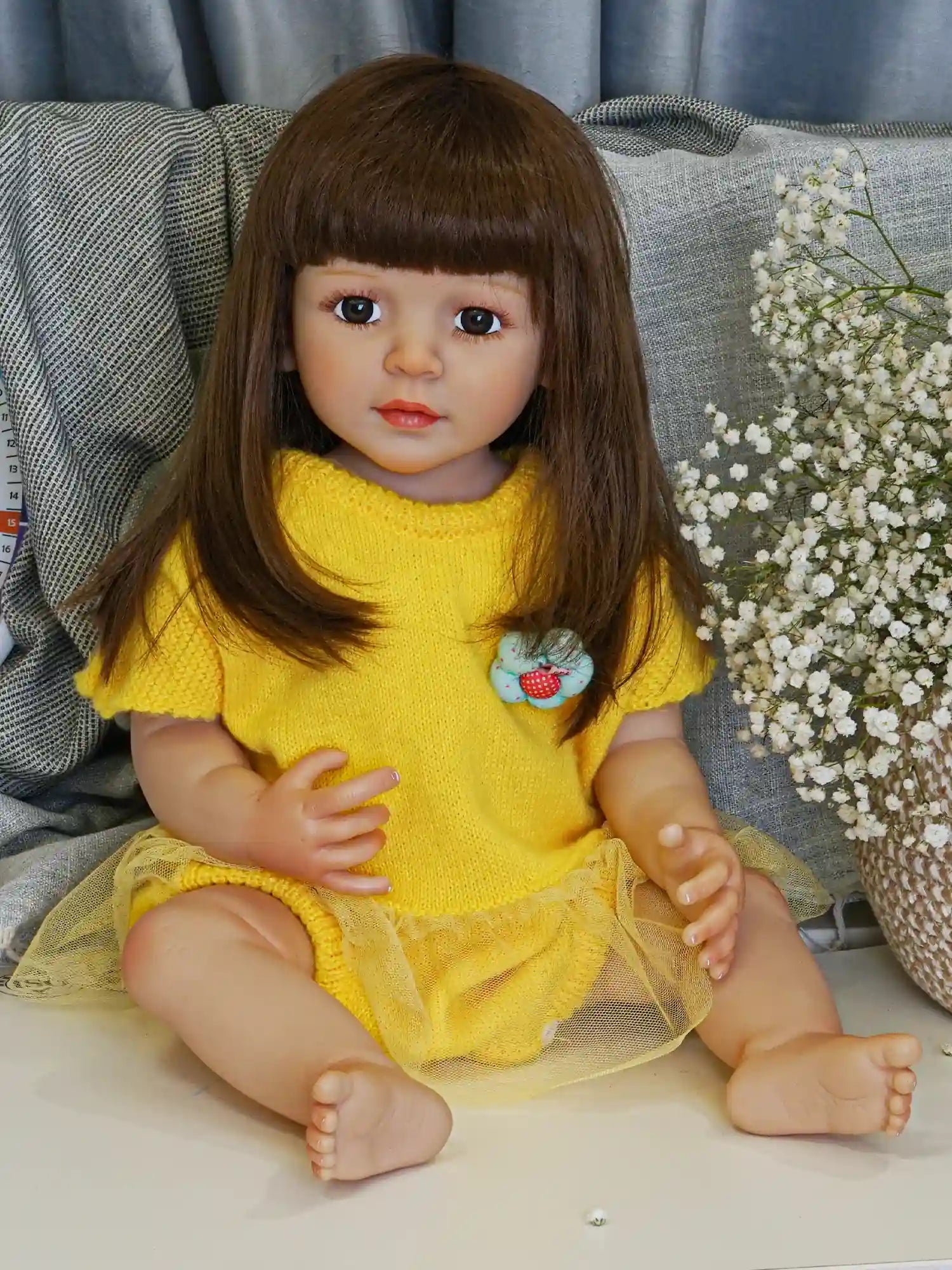 Charming reborn doll seated with a poised posture, wearing a knit yellow dress with a sheer layer, amidst a backdrop of a chic living space with a patterned throw.